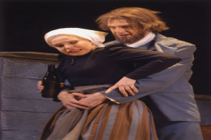 Ein Mann in einem Anzug und eine Frau mit einer Flasche in der Hand stehen nebeneinander auf einer Bühne, mit einer Wand im Hintergrund und Text am unteren Rand des Bildes.