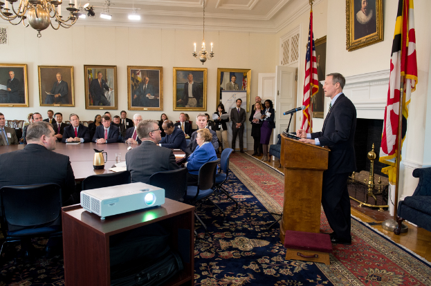 Ein Mann steht an einem Rednerpult und spricht in ein Mikrofon, vor ihm sitzen einige Menschen, links ein Projektor auf einem Tisch, Fotorahmen an der Wand im Hintergrund, rechts zwei Flaggen und Lampen mit einem Kronleuchter oben.
