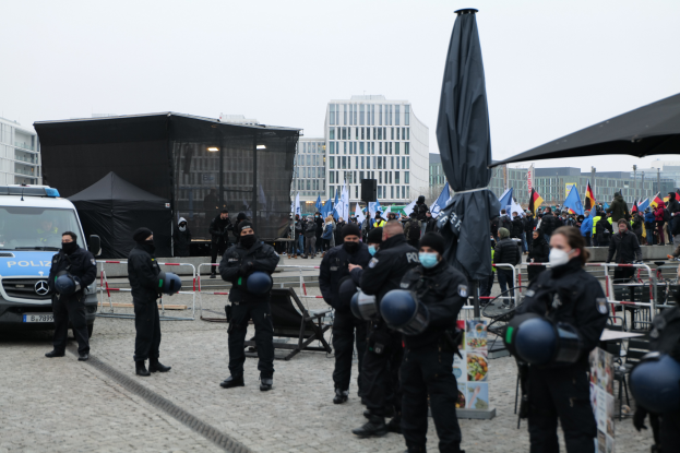 Eine Gruppe von Polizisten steht vor einer Menge von Demonstranten in Berlin, Deutschland, mit Barrikaden, Zelten, Fahnen im Vordergrund und Gebäuden im Hintergrund und einem klaren Himmel.