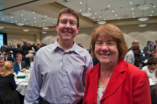 Zwei Personen lachen im Vordergrund, während andere an Tischen mit Tassen und Gläsern sitzen und weitere im Hintergrund an einer Wand diskutieren.
