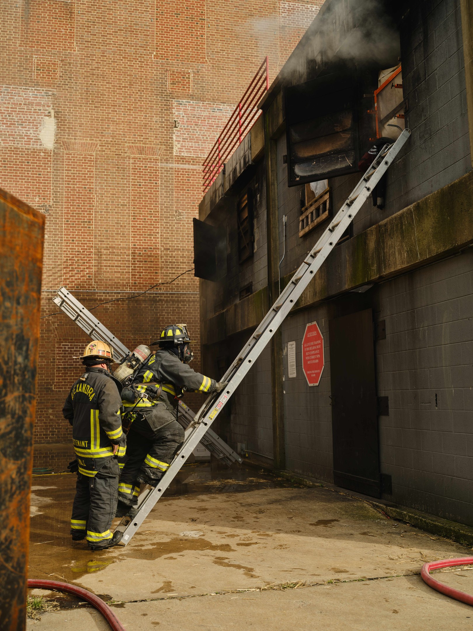Feuerwehrleute mit Helmen, die eine Leiter zu einem rauchgefüllten Gebäude hinaufsteigen, mit einem Metallobjekt links und Rohren an der Basis.