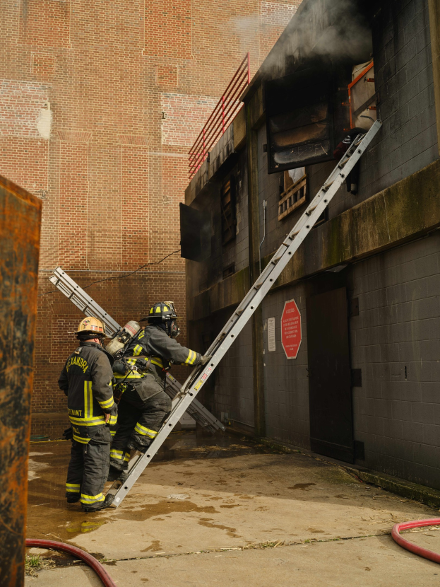 Feuerwehrleute mit Helmen, die eine Leiter zu einem rauchgefüllten Gebäude hinaufsteigen, mit einem Metallobjekt links und Rohren an der Basis.