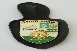 Ein schildförmiges Polizeibadge mit der Aufschrift 'Polizei Berlin' auf einem blauen Hintergrund, einem weißen Rand, einem weißen zentralen Stern und schwarzer fetter Schrift, das auf einer Oberfläche liegt.