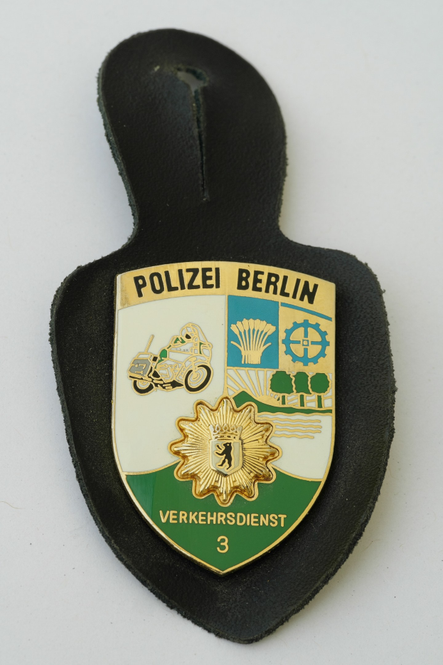 Ein schildförmiges Polizeibadge mit der Aufschrift 'Polizei Berlin' auf einem blauen Hintergrund, einem weißen Rand, einem weißen zentralen Stern und schwarzer fetter Schrift, das auf einer Oberfläche liegt.