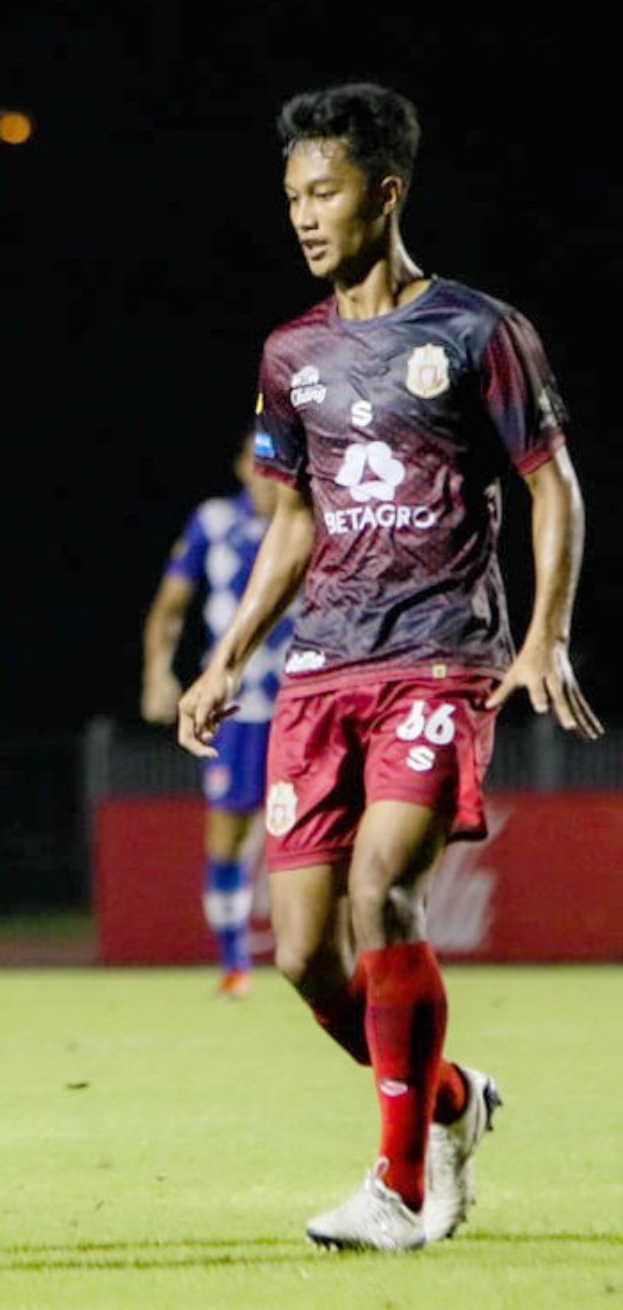 Ein Mann in einem rotbraunen Hemd und roten Shorts spielt Fußball auf einem Feld, mit einer anderen Person im Hintergrund und einer Tafel mit Schrift.