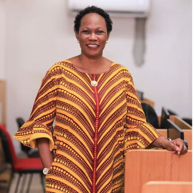 Eine Frau in einem gelben und roten Kleid steht an einem Rednerpult mit einem Mikrofon, spricht in einer öffentlichen Einrichtung mit Stühlen und einer Klimaanlage im Hintergrund.