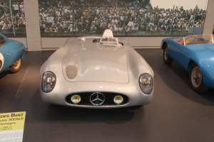 Ein Mercedes-Benz 300 SLR Le Mans Rennauto in einem Museum ausgestellt, umgeben von anderen Autos, mit einer Texttafel links und Menschen, einem Zaun und einer Wand im Hintergrund.