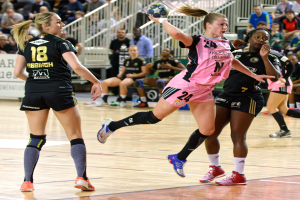 Eine Gruppe von Frauen spielt Handball auf einem Platz mit Zuschauern im Hintergrund.