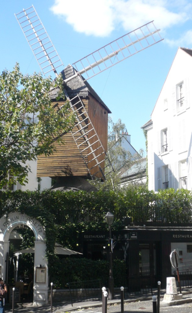 Eine Windm├╝hle, mehrere Geb├Ąude, B├Ąume, Metallstangen und ein Zaun in einer l├╝ndlichen Umgebung.