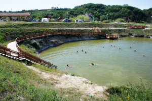 Menschen schwimmen in einem Gewässer umgeben von Grün, mit einer Brücke, Treppen, Schuppen, Fahrzeugen, Pfählen und einem klaren Himmel im Hintergrund.