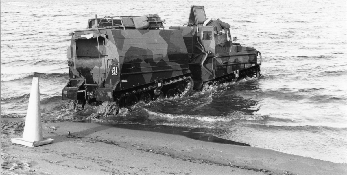 Schwarzes und weißes Foto eines Amphibienfahrzeugs, das durch Wasser fährt, mit einem Verkehrskegel links und Sand am Boden.