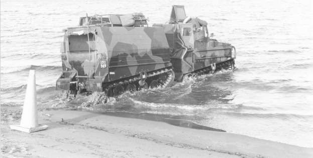 Schwarzes und weißes Foto eines Amphibienfahrzeugs, das durch Wasser fährt, mit einem Verkehrskegel links und Sand am Boden.