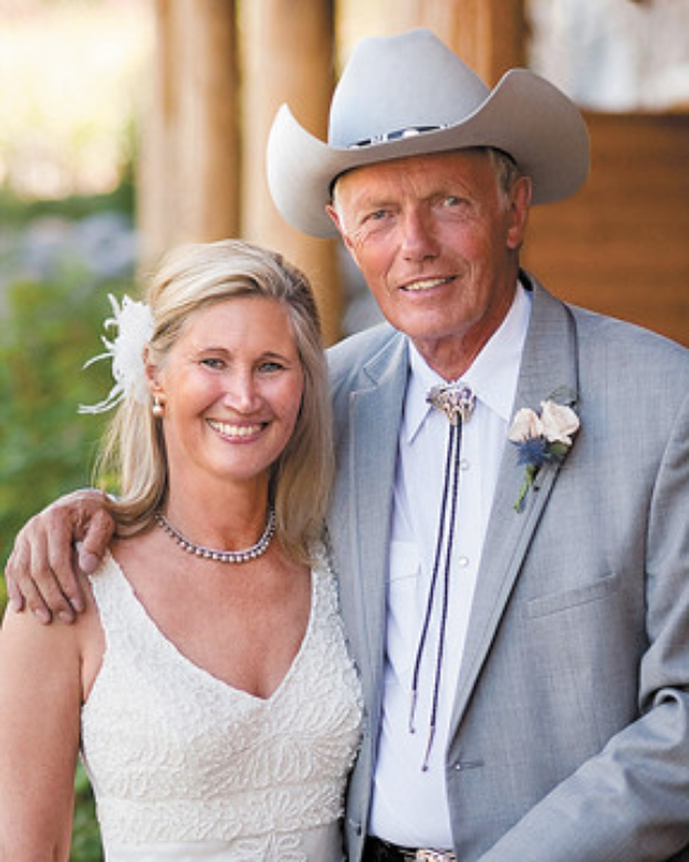 Ein Mann in einem grauen Blazer, weißem Hemd und Hut steht neben einer Frau in Weiß, beide lachend vor einem unscharfen Gebäude, feiern ihren 50. Hochzeitstag.