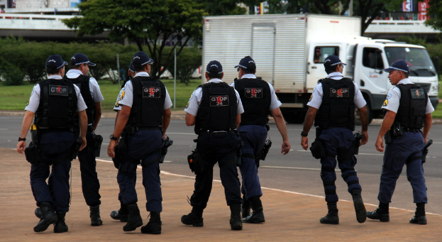 Eine Gruppe von Beamten in Uniform, darunter M√∂tzen, Hemden, kugelsichere Westen, Hosen und Schuhe, geht auf einer Stra√e. Rechts davon steht ein Lastwagen in der N√§he eines Zauns, mit Geb√§uden, B√§umen, Pflanzen, Masten und Bannern im Hintergrund und einer Br√ľcke oben rechts.