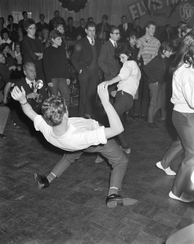 Schwarzes und weißes Bild einer Gruppe von Menschen in Anzügen, die auf einer Tanzfläche vor einem Publikum tanzen, mit einer Wand im Hintergrund, auf der etwas geschrieben steht.