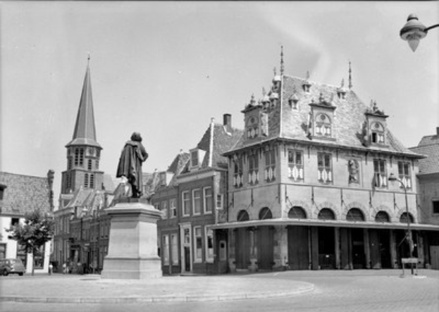 Schwarzes und weißes Foto eines Stadtplatzes mit einer Statue auf einem Sockel in der Mitte, umgeben von Gebäuden, Straßenlaternen, Straßenlaternen, Fahrzeugen auf der Straße, Bäumen und einem klaren Himmel.