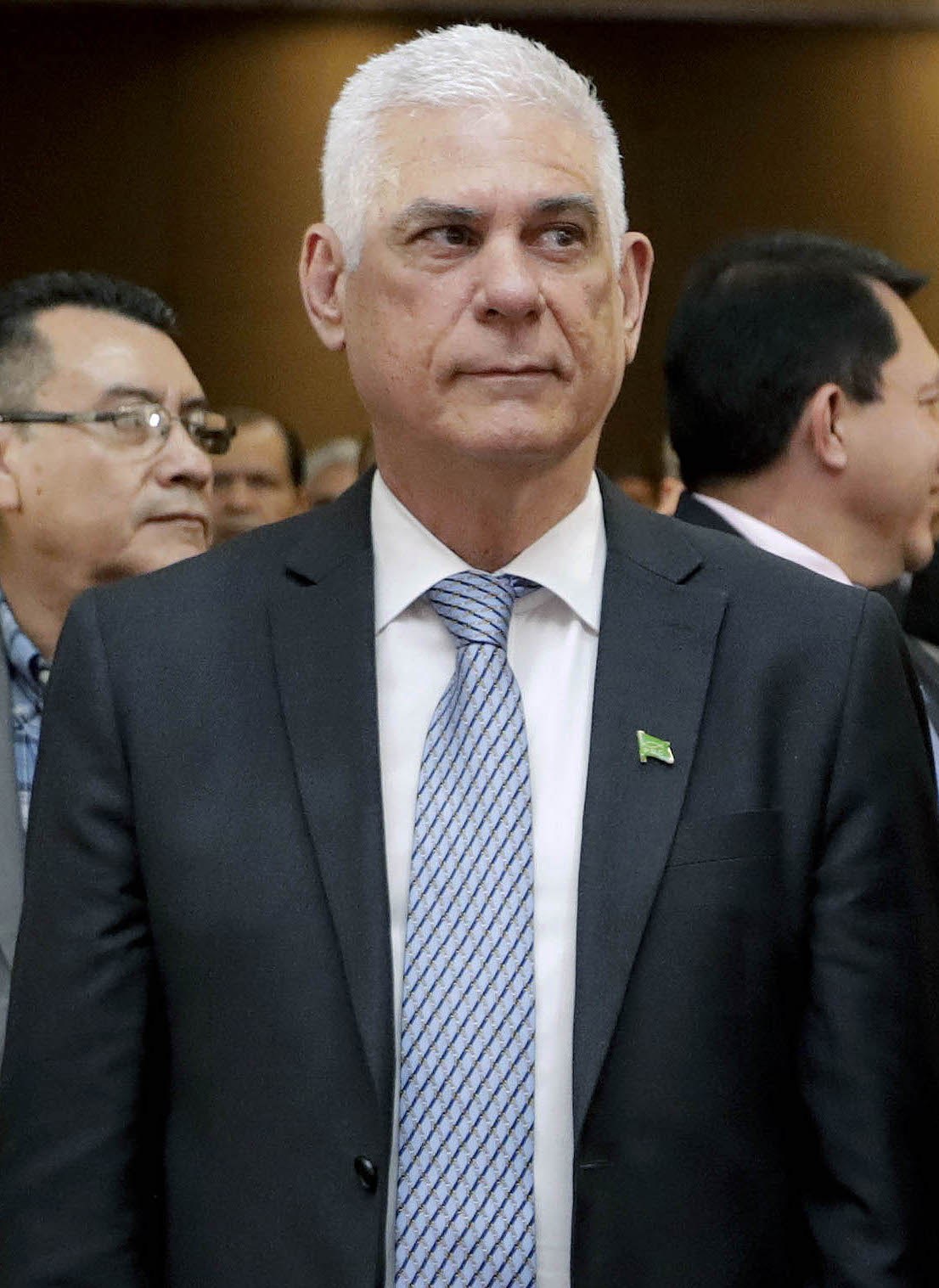 Ein Mann in einem marineblauen Anzug, weißem Hemd und gemusterter Krawatte steht vor einer Gruppe von Menschen mit einer Wand im Hintergrund und hat einen ernsten Gesichtsausdruck.