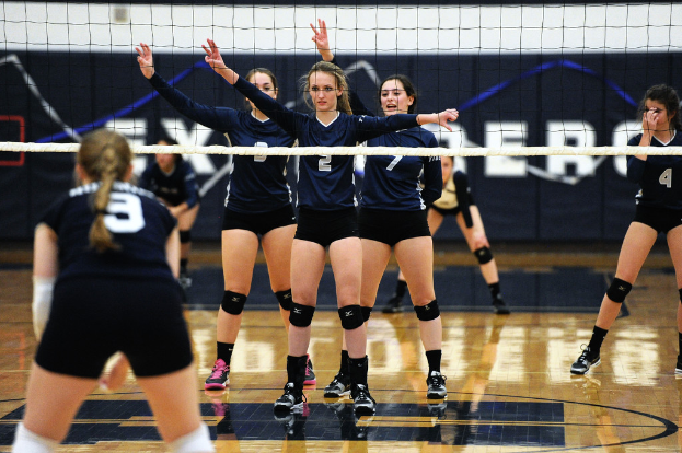 Drei Mädchen bei einem Volleyballspiel auf dem Feld stehend, mit erhobenen Händen, einem Netz vor ihnen und einem Zaun im Hintergrund.