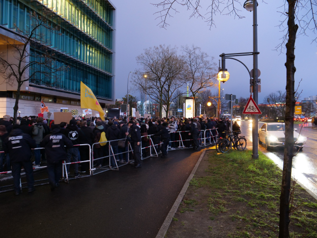 Große Gruppe von Menschen protestiert vor einem Gebäude in Berlin, hält Schilder, mit Barrikaden, Fahrrädern, Laternen, Schildern, Bäumen und Gras im Hintergrund.