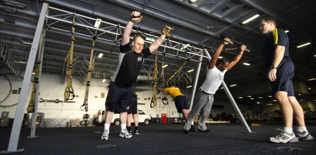 Mannergruppe beim Pull-up-Training in einem Fitnessstudio mit Metallgeräten und Deckenbeleuchtung, mittig ein Mann mit Schuhen.