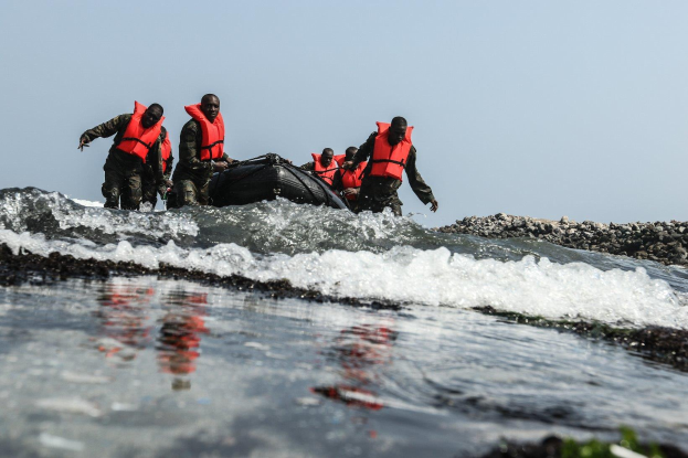 Eine Gruppe von Menschen in Schwimmwesten auf einem Boot im Wasser, umgeben von Felsen auf der rechten Seite und einem klaren Himmel im Hintergrund, scheinen Teil eines Rettungsteams zu sein, das nach Überlebenden sucht.