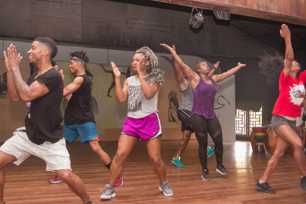 Eine Gruppe von Menschen tanzt in einem Tanzstudio mit einem Gemälde an der Wand, einem Glasfenster auf der rechten Seite und Deckenlampen darüber, was auf eine Zumba-Stunde hinweist.