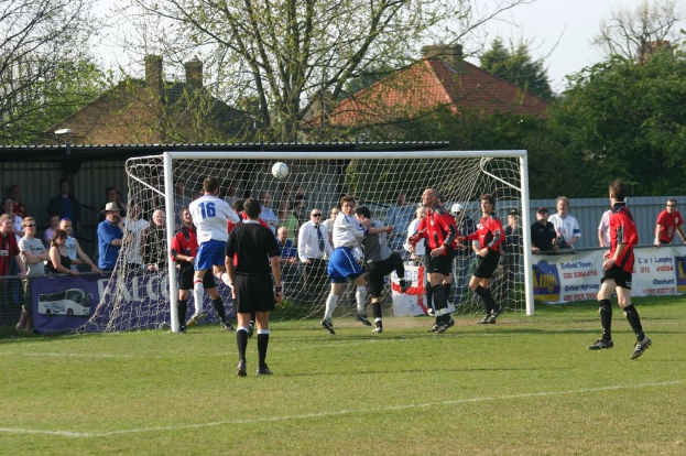 Spieler sind bei einem Fußballspiel auf einem Feld mit einem Tor und Zuschauern im Hintergrund sowie weiteren Bäumen und Häusern im Hintergrund zu sehen.