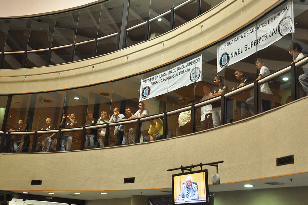 Ein Gebäude mit Menschen davor und zwei Bannern am Glas sowie einem TV-Bildschirm unten im Bild.
