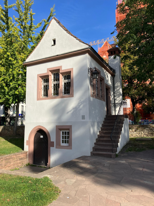 Kleines weißes Gebäude mit schwarzer Tür und Fenstern, das sich in der Altstadt von Heidelberg befindet und eine Treppe, eine Wandlampe, Gras, Bäume, entfernte Gebäude, einen Turm und einen bewölkten Himmel zeigt.