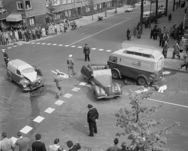 Eine Schwarz-Weiß-Szene eines Autounfalls an einer Straßenkreuzung, mit mehreren Fahrzeugen einschließlich eines Lieferwagens, einer Gruppe von Menschen drumherum und Gebäuden, Bäumen, Polen und Texttafeln im Hintergrund.