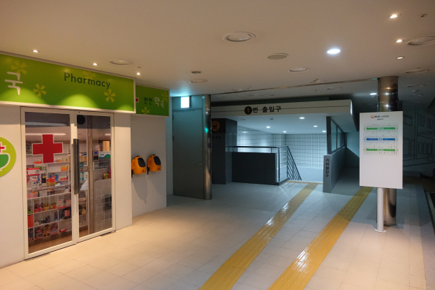 Eingang einer Apotheke mit einem "Apotheke"-Schild links, einer Tafel mit Text rechts, beleuchteten Lampen oben und undeutlichen Hintergrundobjekten.