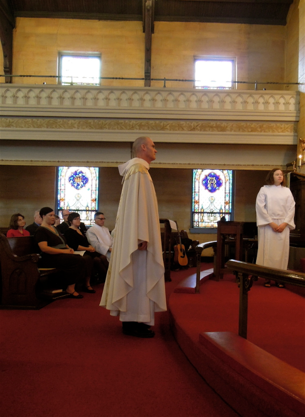 Eine Frau und ein Mann in weißen Kleidern stehen in einer Kirche, mit Menschen auf Bänken links, Musikinstrumenten im Hintergrund und einem roten Teppich, Geländern, Kerzen und Fenstern im Bild.