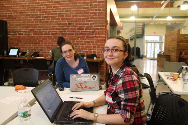 Zwei Frauen an einem Tisch mit Laptops, einer Flasche, einem Stift und Papier in einem Büro mit roter Backsteinwand, Deckenleuchten, einer Treppe und einer Eingangstür im Hintergrund.