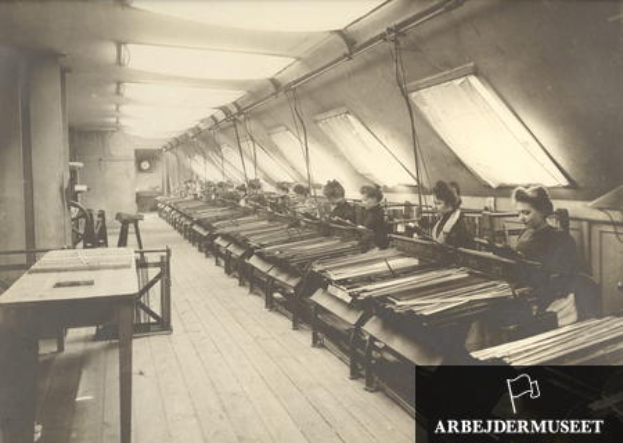 Ein Schwarz-Weiß-Foto von Frauen, die in einer Textilfabrik an Maschinen arbeiten, die an Tischen auf der linken Seite sitzen, ein Uhr an der Wand im Hintergrund und Text in der rechten unteren Ecke.