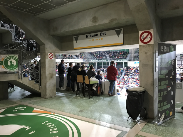 Gruppe von Menschen vor einer sitzenden Menge in einem Stadion mit Schildern 'Boston Celtics vs New York Jets', einem Müllbehälter, einer Tafel mit Text, einer Treppe mit Geländern und einem Dach mit Deckenleuchten.