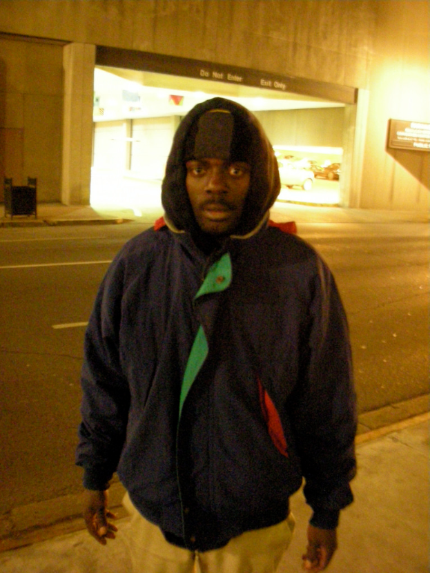 Eine Person in einer Jacke steht mit dem Rücken zur Kamera, vor einer Wand und einer Straße.