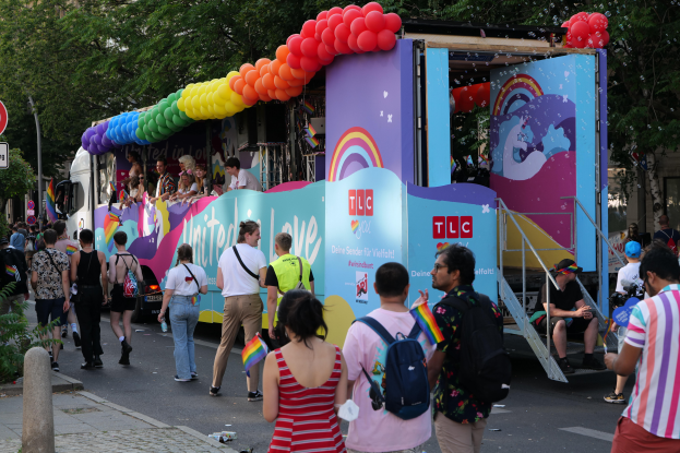 Eine Gruppe von Menschen geht eine Straße entlang neben einem Lastwagen mit bunten Luftballons, mit Schildern an Pfosten an der Straße und Bäumen und Gebäuden im Hintergrund, was auf eine Pride-Parade in Paris hindeutet.