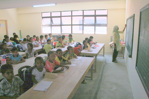 Ein Klassenzimmer mit einer Frau, die auf einer Tafel rechts schreibt, Fenstern in der Mitte und einer Lampe an der Decke, mit Tischen voller Bücher und Wasserflaschen sowie Stühlen mit sitzenden Kindern.