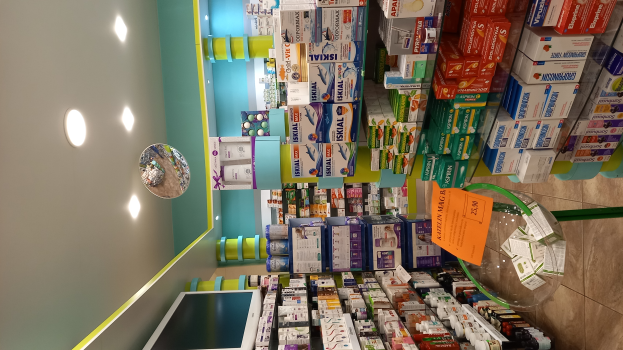 Eine Apotheken-Theke mit verschiedenen Medikamenten in Boxen und Flaschen auf Gestellen, mit einem Spiegel an der Wand, Deckenleuchten und einem Glastisch mit Papieren und Gegenständen darauf, der Boden ist unten zu sehen.