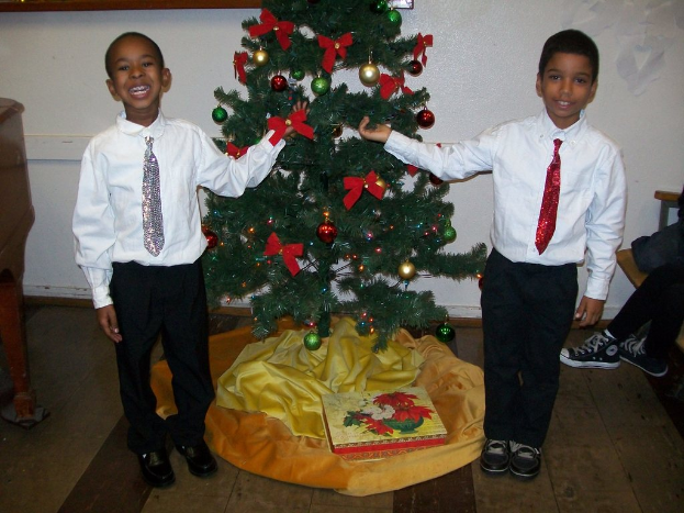 Ein geschmückter Weihnachtsbaum steht in einem Raum mit einer Kiste und anderen Gegenständen auf dem Boden, einem hölzernen Tisch auf der linken Seite, einem Stuhl auf der rechten Seite und einer weißen Wand mit angebrachten Gegenständen im Hintergrund; zwei Jungen in weißen Hemden lächeln, während sie den Baum halten.