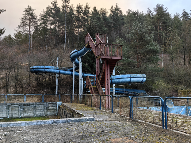 Ein verlassener Wasserpark mit einer blauen Wasserrutsche in der Mitte, umgeben von Geländern, Treppen und einer Wand, mit Bäumen und einem klaren blauen Himmel im Hintergrund und Text am unteren Rand des Bildes.