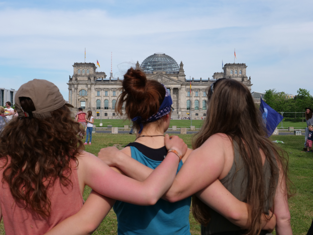 Drei Frauen in verschiedenen farbigen Kleidern, eine trägt eine Mütze, stehen vor dem Reichstaggebäude in Berlin, Deutschland, mit Menschen, Flaggen, Bäumen, Wolken und dem Himmel im Hintergrund.