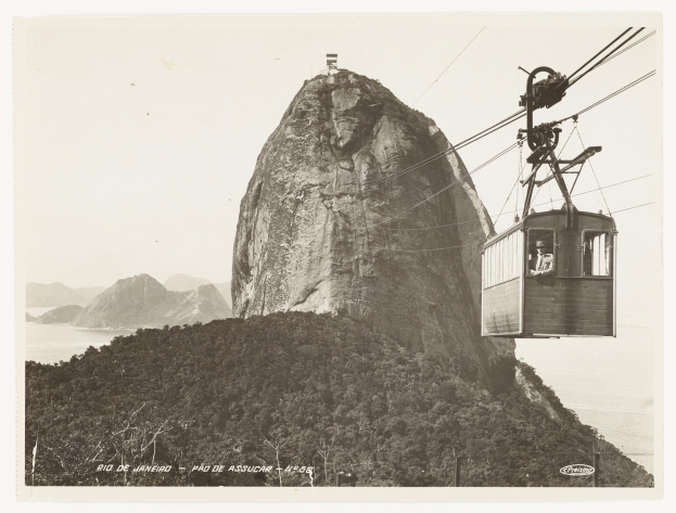 Schwarzes und weißes Foto einer Seilbahn, die einen Berg hinauffährt, mit einer Person darin, umgeben von Bäumen, Hügeln und Wasser, und Text unten.