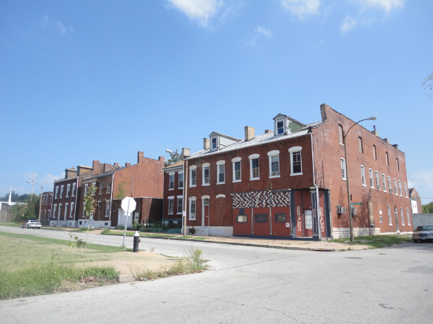 Ein Gebäude mit Pfählen und Straßenlaternen, umgeben von Bäumen und Fahrzeugen auf beiden Seiten, mit einem Weg, Gras und einem Feuerlöscher unten und Himmel oben.