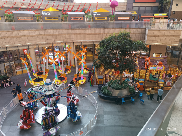 Ein Vergnügungspark in einem Einkaufszentrum mit verschiedenen Attraktionen, Bäumen, Menschen, Stühlen, Ständen und Gebäuden, mit einem Wasserzeichen in der rechten unteren Ecke.