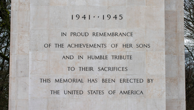 Ein Denkmal mit einer Tafel, auf der "In stolzem Gedenken an die Taten ihrer Söhne und in Demut zu ihrem Opfer" steht, vor einem Hintergrund aus Bäumen und einem klaren blauen Himmel.