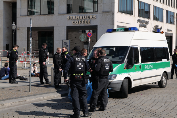 Polizisten vor einem Starbucks-Café mit einem Van auf der rechten Seite und Menschen auf der linken Seite, mit einem Gebäude, einem Schild, einer Straßenlaterne und einem Zaun im Hintergrund.