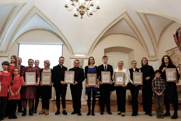 Gruppe von Menschen mit Urkunden bei einer Preisverleihung mit sichtbarer Hand, Wand, Tür, Bildschirm und Kronleuchter im Hintergrund.