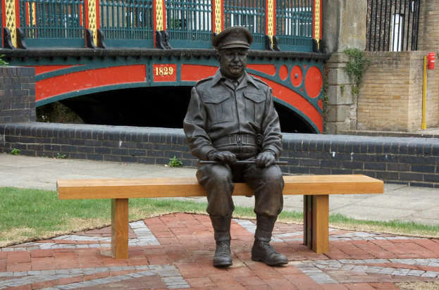 Eine eiserne Statue eines Mannes sitzt auf einer Pappbank, mit einer brückenähnlichen Struktur dahinter, die das Jahr 1829 in Gold anzeigt, flankiert von schwarzen Backsteinmauern und Gittern.