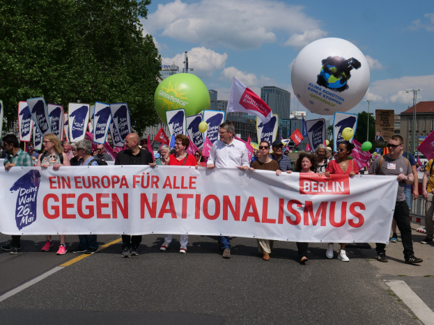 Gruppe von Menschen bei einer Demonstration gegen Nationalismus in Berlin, die eine Fahne, Flaggen und Luftballons halten, mit Bäumen, Gebäuden und einem bewölktem Himmel im Hintergrund.